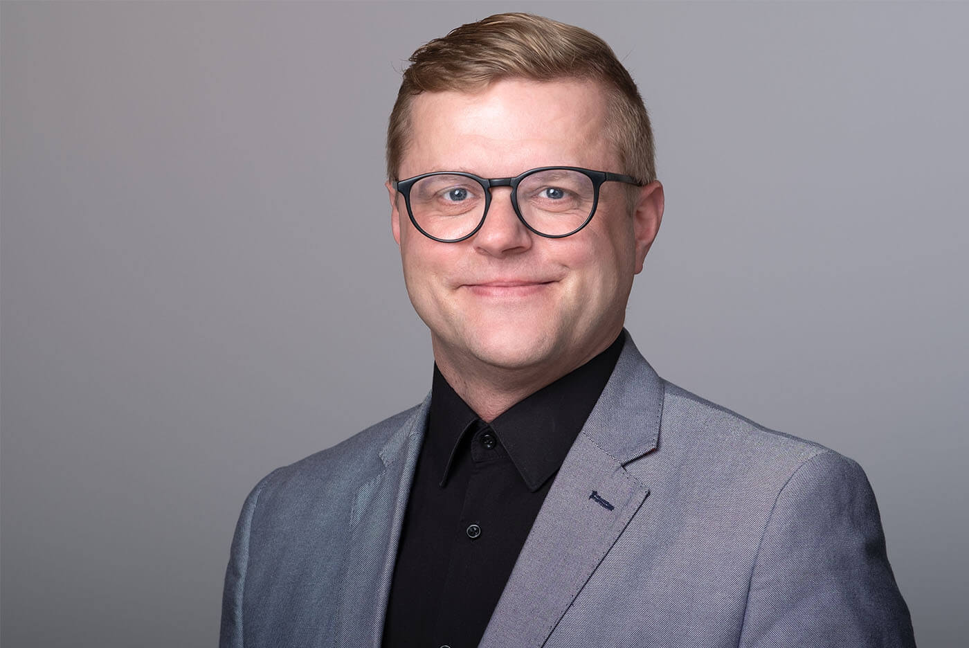 Bewerbungsfoto eines Mannes mit schwarzem Hemd, Brille und Sakko vor grauem Hintergrund, aufgenommen im Fotostudio KARRIEREPORTRAITS in Dortmund